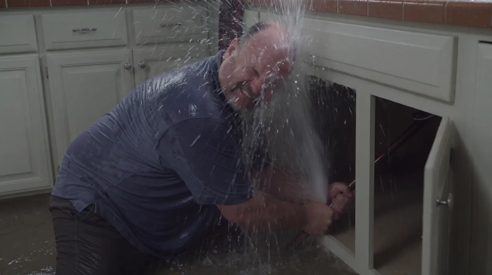 A plumber crouched at an open kitchen cabinet taking a jet of water full in the face from the pipe he is holding, the floor already soaked.